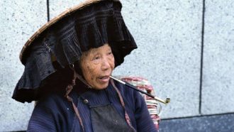 2E9R66G Hong Kong, China. A Hakka woman smoking a pipe outside a temple in the New Territories wearing a traditional hat.