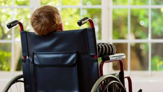 Backview of a  9 year old blond boy sitting in a wheelchair looking out of a window at a garden he cannot reach or play in.