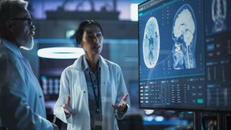 Neuroscience Laboratory Meeting: Female Asian Scientist And Male Caucasian Surgeon Discussing MRI Brain Scan On TV Display. Specialists Working On Medication For Rehabilitation After Brain Damage.